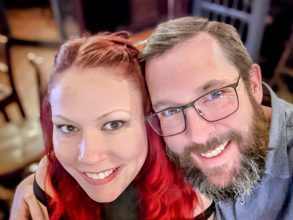 Close up portrait of a vibrantly redheaded woman and bearded man in glasses, both grinning.